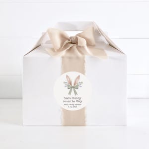 May include: A white gift box with a beige ribbon and bow. A round sticker with a bunny illustration and the text "Some Bunny is on the Way" is attached to the box. The text "Anna's Baby Shower 4.12.2025" is also on the sticker.