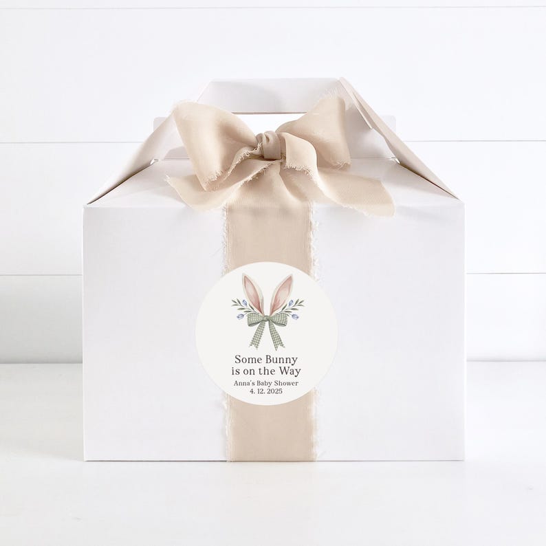 May include: A white gift box with a beige ribbon and bow. A round sticker with a bunny illustration and the text "Some Bunny is on the Way" is attached to the box. The text "Anna's Baby Shower 4.12.2025" is also on the sticker.