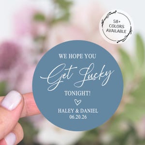 May include: A round, dusty blue sticker with the text "We Hope You Get Lucky Tonight!" in white script. Below the text is a heart symbol, followed by names and a date. A smaller circle indicates 58+ colors are available.