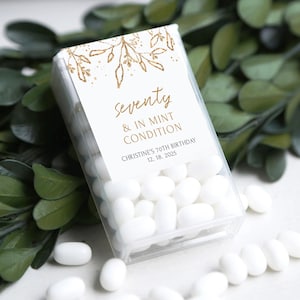 May include: A clear plastic box filled with white mints. The box has a white label with gold text that reads "Seventy & In Mint Condition" and "Christine's 70th Birthday 12.18.2025". Gold floral design at the top.