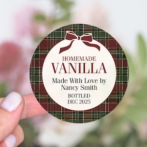 May include: Round label with a plaid pattern in green, burgundy, and gold. The label reads "Homemade Vanilla" with the text "Made With Love by Nancy Smith" and "Bottled Dec 2025". A burgundy bow is at the top.