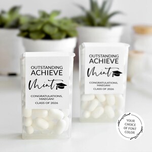 May include: Two transparent plastic containers filled with white mints. Each has a white label with the words "OUTSTANDING ACHIEVE Mint" and "CONGRATULATIONS, MAEGAN! CLASS OF 2026" printed in black. A graduation cap graphic is also included.