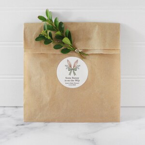 May include: A brown paper bag with a white circle sticker. The sticker has a cartoon bunny with a bow and the text "Some Bunny is on the Way" with the date "4.12.2023".