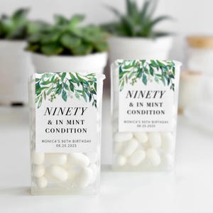 May include: Two clear plastic containers filled with white mints. Each container has a label with the text "NINETY & IN MINT CONDITION" and "MONICA'S 90TH BIRTHDAY 08.20.2025". The labels feature a green leaf design.