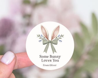 May include: A round sticker with a bunny ears design, a green plaid bow, and the text "Some Bunny Loves You From Oliver". The bunny ears are pink and the bow is green and white. The sticker has a floral design.