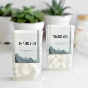 May include: Two clear plastic containers filled with white sweets. Each container has a label that reads "THANK YOU" above the text "JENNIFER'S BACHELORETTE 08.20.2025". The label features a watercolour mountain range design in grey and blue.