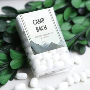 May include: A transparent plastic box filled with white sweets. The box features a label with the text "CAMP BACH", "JENNIFER'S BACHELORETTE 08.20.2025", and a mountain design. The sweets are scattered around the box.