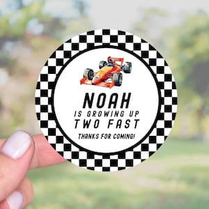 May include: A round sticker with a black and white checkered border. The center features a red race car illustration and the text "NOAH IS GROWING UP TWO FAST THANKS FOR COMING!" on a white background. The sticker is held by a hand.