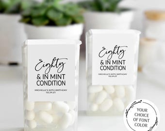 Puede incluir: Dos recipientes de plástico transparente llenos de mentas blancas. Cada recipiente tiene una etiqueta blanca con el texto "Eighty & IN MINT CONDITION" y "MICHELLE'S 80TH BIRTHDAY 02.24.25" en escritura negra. Un gráfico circular dice "YOUR CHOICE OF FONT COLOR".