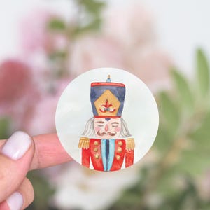 May include: A round sticker with a watercolor illustration of a nutcracker. The nutcracker is wearing a red and blue uniform with gold accents. The sticker has a white background.