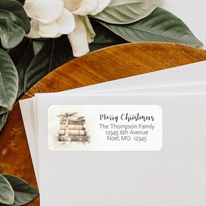 May include: A white rectangular return address label with a watercolour illustration of a stack of books with a ribbon and pine branches. The text "Merry Christmas" is at the top of the label, followed by "The Thompson Family", "12345 6th Avenue", and "Noel, MO 12345".