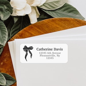 May include: White rectangular label with a watercolor-style black bow illustration. The label includes the name "Catherine Davis" and an address: 12345 6th Avenue, Pleasantville, Ny 12345. The label is on a stack of white paper.