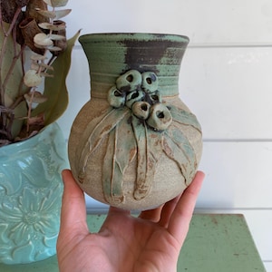 May include: A round ceramic vase with a tan, textured body and a green-glazed rim. The exterior features a raised, sculpted botanical design of eucalyptus leaves and gum nuts in shades of green and brown, applied to the front of the vessel.