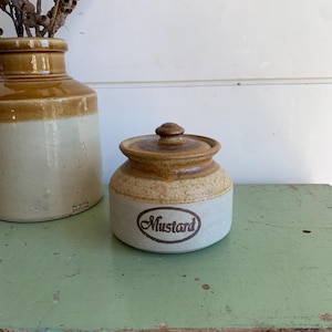 May include: A small, round ceramic mustard pot with a lid, featuring a two-tone design with a speckled tan upper section and a matte off-white base. The word Mustard is printed in a dark brown script font inside an oval frame on the front.