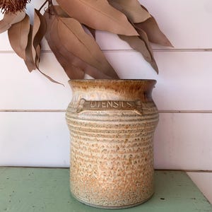 May include: A cylindrical ceramic utensil crock featuring a speckled tan and brown glaze with a darker rim. The word UTENSILS is embossed on a raised rectangular plaque on the upper front section of the vessel, which has a slightly flared top edge.