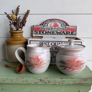 May include: A Port Curtis Pottery stoneware creamer and sugar bowl set with a white glaze and hand-painted pink floral motifs. The set includes a lidded sugar bowl and a creamer, both featuring pink rim accents, displayed in front of the original box.
