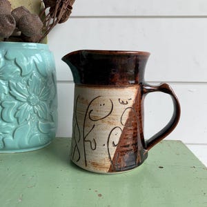 May include: A ceramic pitcher featuring a two-tone glaze with a dark brown upper section and a cream-coloured lower body. The lower portion is decorated with hand-drawn black line art depicting abstract faces and shapes, complemented by a handle.