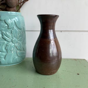 May include: A dark brown ceramic vase with a smooth, glossy finish and a narrow neck. The vase features a subtle embossed circular pattern around the upper body and a small, circular maker's mark stamped near the base on the front side.