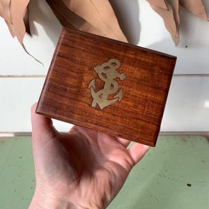 May include: A rectangular wooden box with a polished brown finish featuring a centred brass inlay of a nautical anchor. The box has a smooth, grained surface and a hinged lid design, showcasing a classic maritime aesthetic with metallic detailing.