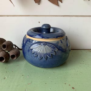 May include: A round, blue glazed ceramic sugar bowl with a matching lid. The exterior features a raised, embossed floral or leaf-like pattern around the centre. The base of the bowl shows a natural, unglazed tan clay rim, and the lid has a knob.
