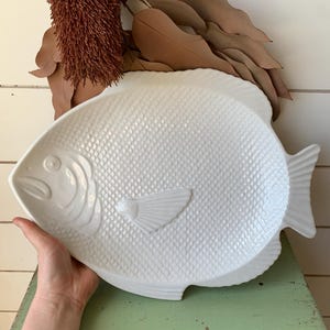 May include: A white ceramic serving platter shaped like a fish, featuring a detailed embossed scale pattern across the body, a smooth head section with a defined eye and mouth, and a textured fin and tail design on a glossy, solid white finish.