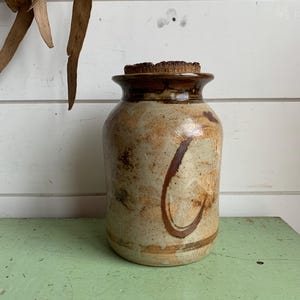 May include: A handmade ceramic jar with a cork stopper. The jar features a beige base with brown and orange accents and a large brown 'C' design. The jar is approximately 18 cm tall and 13 cm wide, suitable for kitchen or home decor.