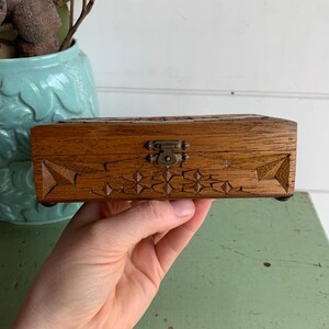 May include: A rectangular wooden box featuring a dark brown finish with intricate geometric carvings on the front panel. The box is secured with a small brass-coloured metal latch centred on the front edge and rests on small, rounded feet.