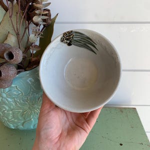 May include: A round, off-white ceramic bowl featuring a hand-painted design of green eucalyptus leaves and brown gum nuts on the inner rim. The bowl has a smooth, matte finish and a simple, circular shape, showcasing a natural botanical motif.