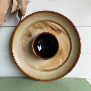 May include: A round ceramic serving dish with a built-in dip bowl. The dish has a light beige centre with brown and tan accents and a dark brown rim. The dip bowl is dark brown. The dish is on a green surface.