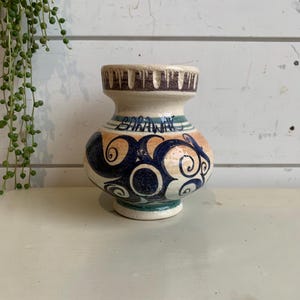 May include: A small, white ceramic vase with blue and orange painted swirls and the word "Baranya" written on the side.