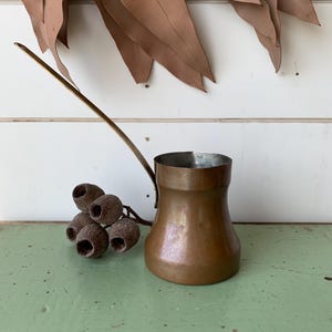 May include: A copper coffee pot with a long, slender brass handle and a wide, rounded base. The interior of the pot is lined with a silver-toned metal. The exterior surface shows a warm, metallic patina with subtle variations in colour.