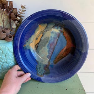 May include: A large, round ceramic bowl featuring a deep cobalt blue glaze with a wide, smooth rim. The interior surface displays a decorative, abstract blend of earthy tones, including streaks of golden yellow, muted green, and burnt orange.