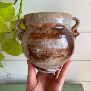 May include: A round, speckled ceramic pot with two small, decorative side handles. The glaze features a blend of light beige, tan, and deep brown tones with a glossy finish. The vessel has a wide opening and a tapered base, showcasing a rustic, hand-crafted appearance with visible colour variations across the surface.
