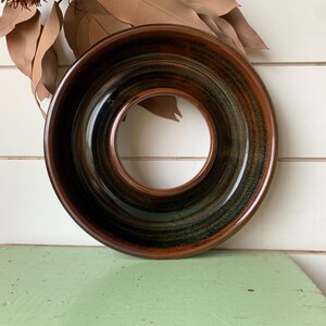 May include: A circular ceramic vessel with a wide, flat rim and a large central opening. The piece features a glossy glaze with blended tones of deep brown, black, and hints of dark green, creating a smooth, reflective surface throughout the object.