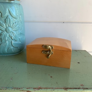 May include: A small, rectangular wooden box with a smooth, light brown finish and a curved lid. The box features a central brass-coloured metal latch mechanism on the front, secured with small screws, designed to keep the lid closed securely.