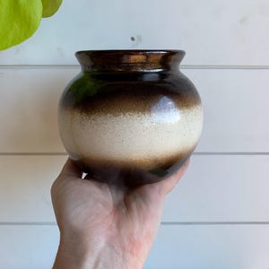 May include: A small, round ceramic vase with a brown and white gradient. The vase has a dark brown rim and a smooth, glossy finish. The vase is held in a hand against a white background.