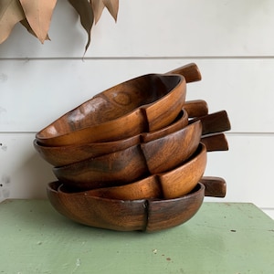 May include: A stack of six wooden serving bowls with attached handles. The bowls are oval-shaped and made of dark brown wood. The bowls are stacked on top of each other, with the handles facing outwards. The bowls are on a green surface.