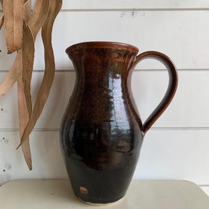 May include: A handmade, dark brown ceramic jug with a handle. The jug has a glossy finish with colour variations from light brown to nearly black. The jug is likely used for serving beverages or as a decorative item.