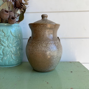 May include: A brown ceramic jar with a speckled finish, a rounded body, and a matching lid with a small knob handle. The jar features two small side loops and a smooth, glazed surface with subtle variations in tone from light to dark brown.