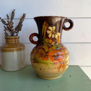 May include: A ceramic vase with two side handles, featuring a gradient from dark brown at the top to a light beige base. The body is ribbed and decorated with hand-painted yellow and orange flowers. Beside it sits a smaller, two-toned brown jar.