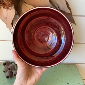 May include: A round ceramic bowl with a deep, glossy burgundy red glaze featuring a subtle spiral pattern etched into the interior surface. The bowl has a clean, white rim that contrasts with the dark red finish, creating a distinct circular border.