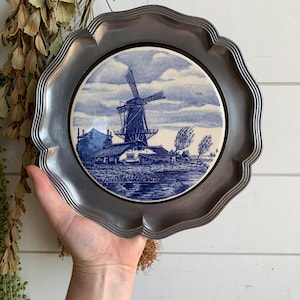 May include: A decorative plate featuring a blue and white windmill scene. The plate has a scalloped, silver-coloured metal rim. The windmill scene is centered on the plate and depicts a windmill, water, and a cloudy sky.