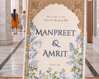May include: A wooden easel displays a welcome sign for a wedding. The sign features ornate gold detailing and floral accents in blue and white. The text reads "Welcome to the Anand Karaj of Manpreet & Amrit 26.07.26."