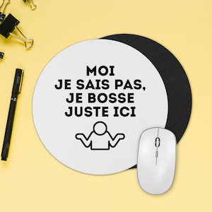 Peut inclure: Un tapis de souris rond blanc avec le texte "MOI JE SAIS PAS, JE BOSSE JUSTE ICI" en noir. Une illustration en noir et blanc d'une personne haussant les épaules est en dessous du texte. Une souris d'ordinateur blanche est à droite.