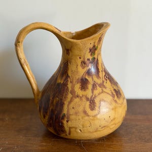 May include: A ceramic pitcher with a rounded body and a curved handle. The surface features a mustard yellow base glaze with mottled, dark brown and subtle green drip patterns throughout the exterior, creating a textured and variegated appearance.
