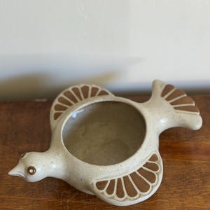 May include: A ceramic bird-shaped bowl with a speckled off-white finish. The piece features brown painted details on the wings and tail, with a circular opening in the center of the body. The bird has a small head with a pointed beak and eye.