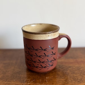 May include: A ceramic mug with a matte terracotta red body featuring a repeating pattern of small black dragonflies. The upper rim and interior are finished in a speckled beige glaze, and the mug includes a matching terracotta red handle.