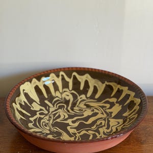 May include: A shallow round ceramic bowl featuring a dark brown interior glaze with a swirling cream-colored marbled pattern. The exterior of the bowl is a matte terracotta color, and the rim has a textured, ridged edge detail throughout.