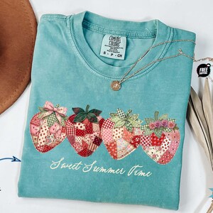 May include: A seafoam green short-sleeve t-shirt featuring a graphic of four strawberries with a patchwork quilt pattern in shades of red, pink, and white. The text "Sweet Summer Time" is printed in white script below the strawberries.