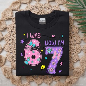 May include: A black t-shirt featuring a graphic design with the text I WAS NOW I'M and the numbers 6 and 7. The number 6 is pink with floral patterns and blue bows, while the number 7 is purple with floral patterns and a pink bow, surrounded by small icons.